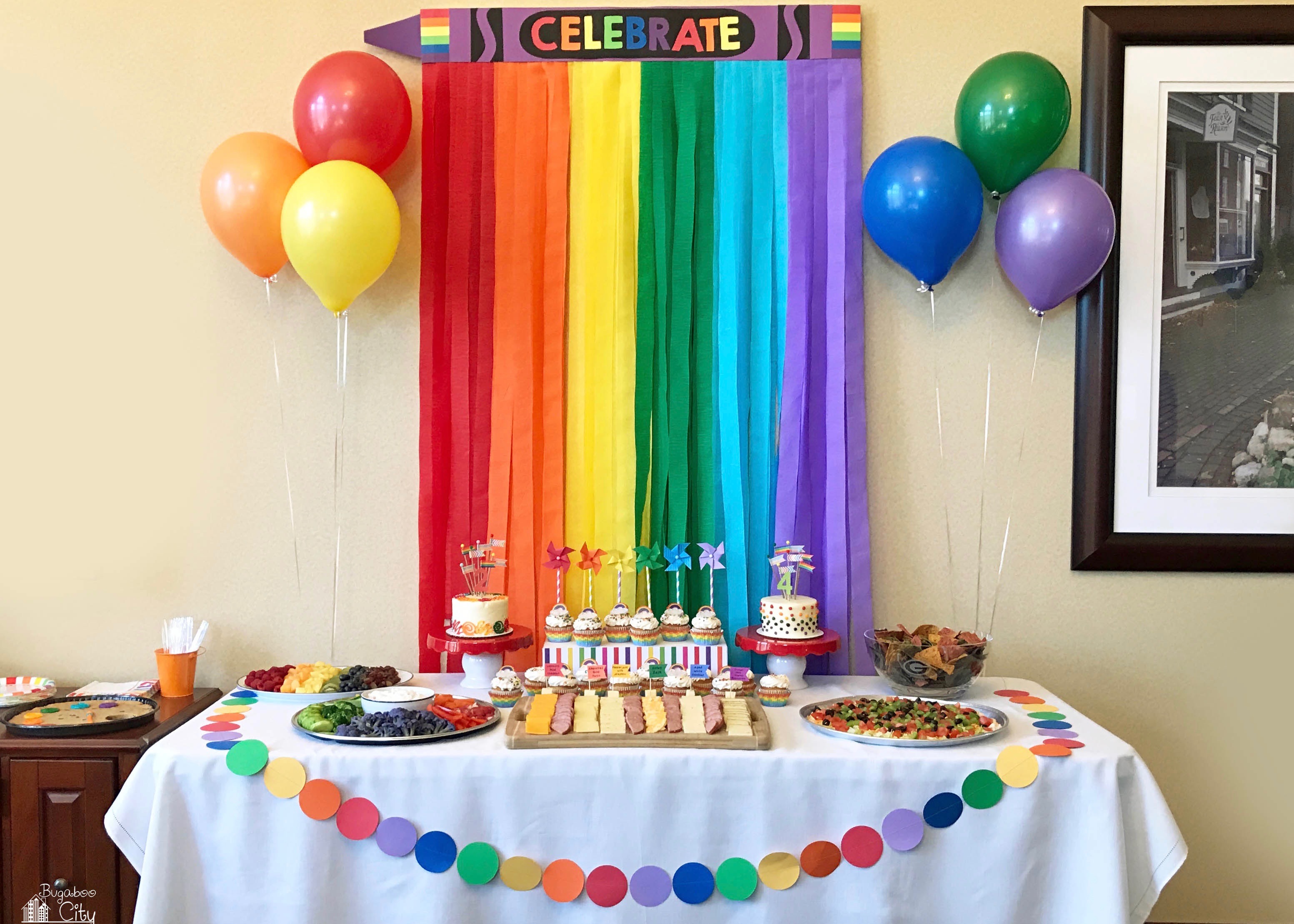  A rainbow of streamers serve as backdrop for a birthday party decorated in rainbow theme with personalized touches like a name banner, rainbow balloons, and a cake with rainbow sprinkles.
