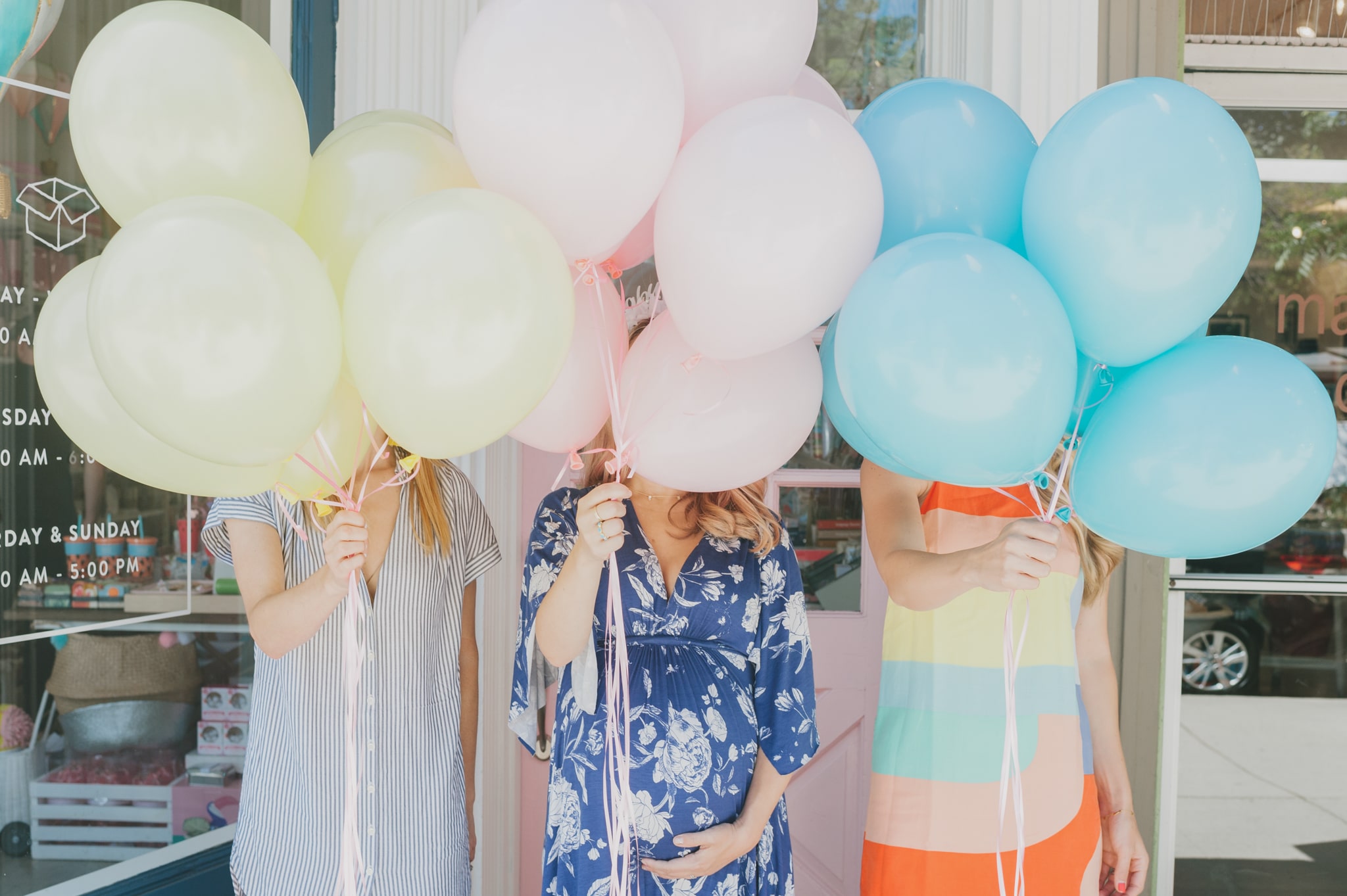 Three girls holding yellow, pink and blue bunches of balloons in front of their faces - Project Nursery